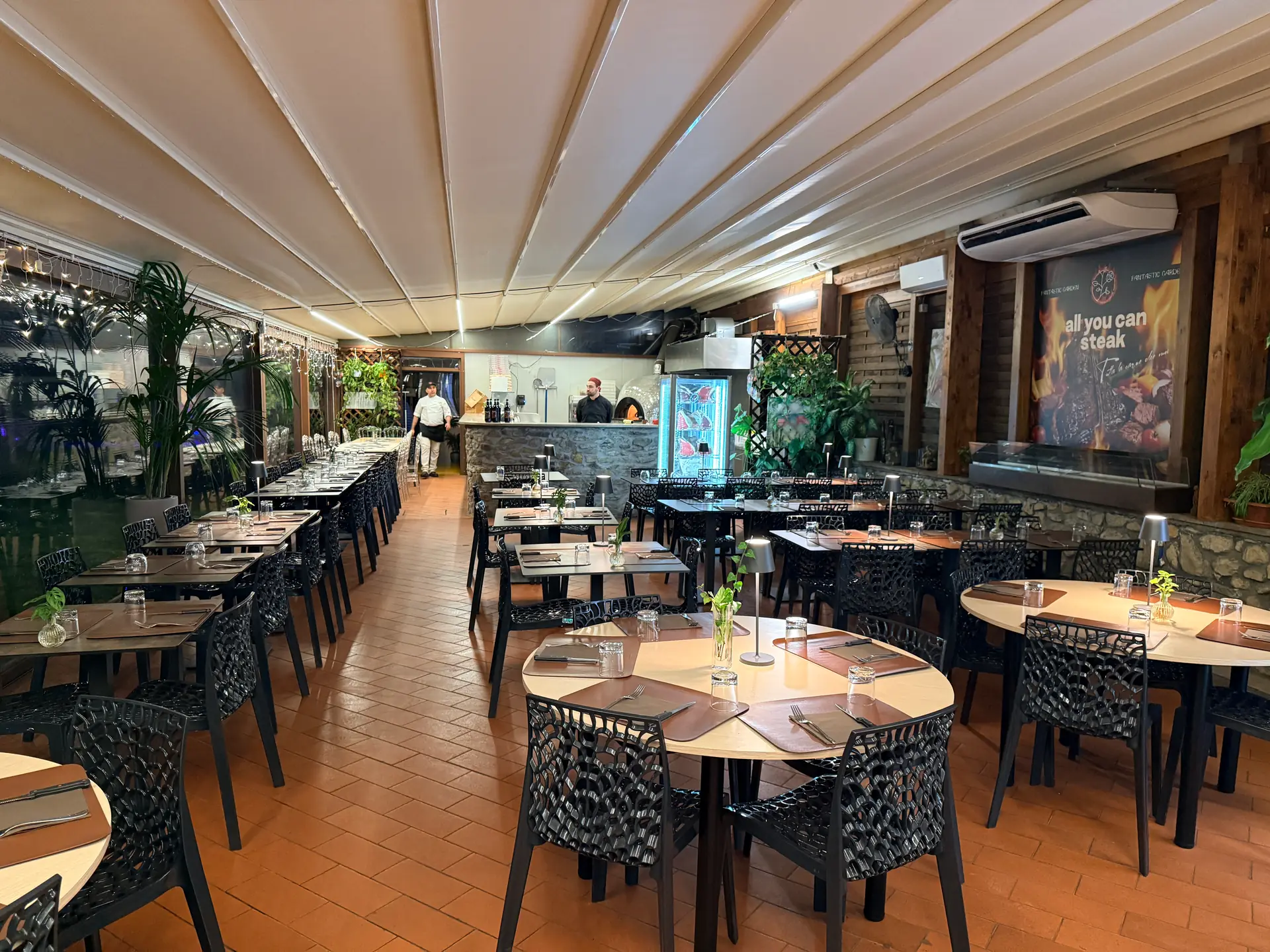 The dining room at All You Can Steak in Sesto Fiorentino: tables laid under the veranda, the wood-fired oven at the back and the team at work before the evening service